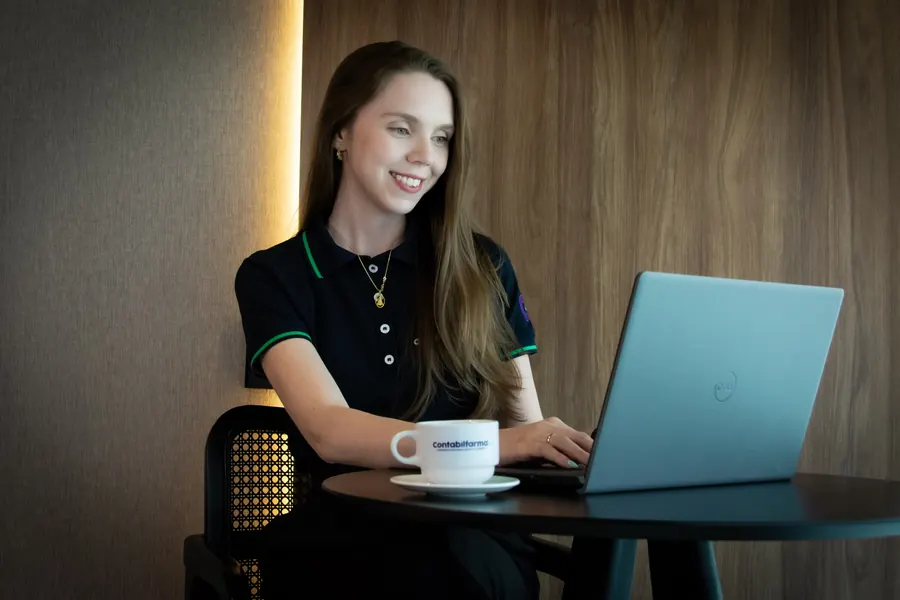 Colaboradora sorrindo enquanto trabalha no notebook, com uma xícara da Contabilfarma na mesa