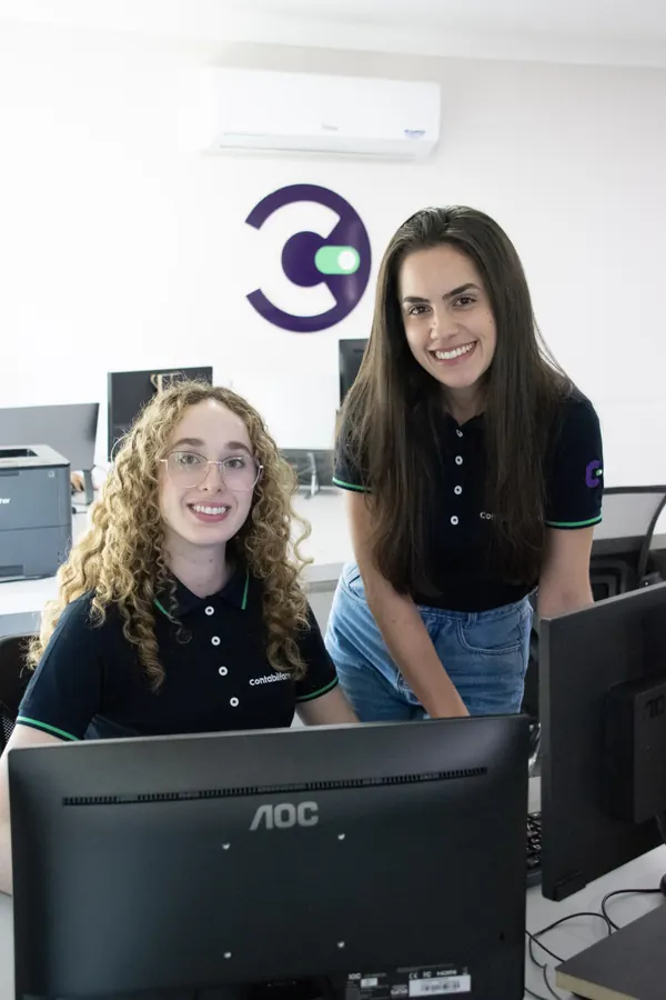 Duas colaboradoras sorrindo em frente ao símbolo da Contabilfarma na parede do escritório